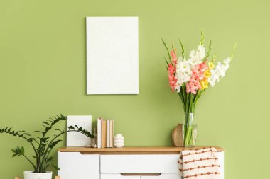 Kitaplar, biblo ve vazo, oturma odasındaki yeşil duvarın yakınındaki çekmecelerde bulunan gladyatör çiçekleri.