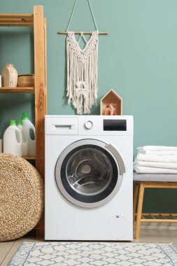 Modern çamaşır makinesi, pouf ve raf ünitesi banyonun içindeki yeşil duvarın yanında, yakın plan.