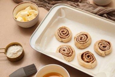 Çiğ tarçınlı çörek tepsisi ve bej arka planda malzemeler, yakın plan.