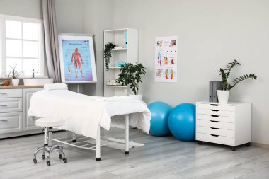 Modern tıp merkezinde kanepe, spor topu ve posterli masaj terapisti ofisi.