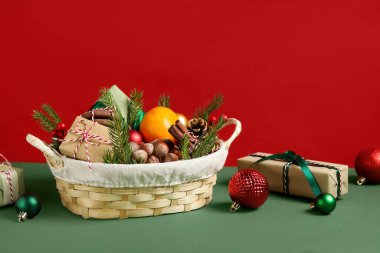 Kırmızı duvarın yanındaki yeşil masada, içinde ürünler ve Noel topları olan hediye sepeti.