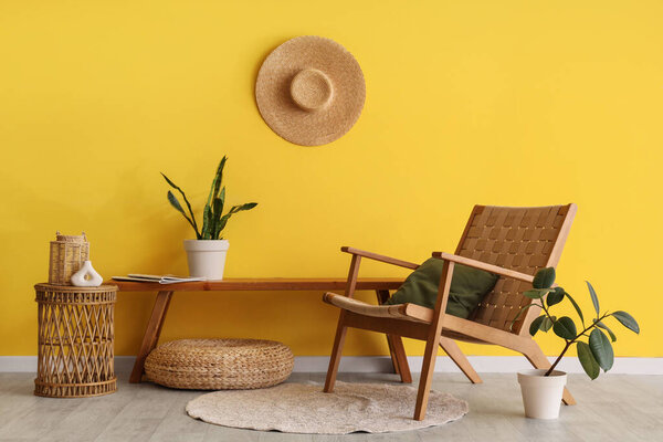 Wooden bench with armchair, wicker table and houseplants near yellow wall in room