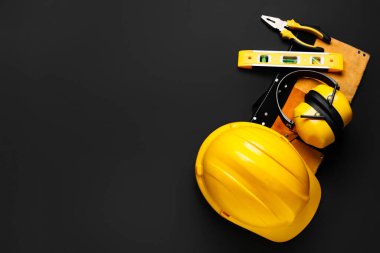 Sert şapka, koruyucu kulaklıklar, alet kemeri ve siyah arka planda inşaat araçları, üst görünüm