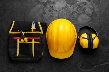 Sert şapka, alet çantası ve siyah grunge arka planda koruyucu kulaklık, üst görünüm