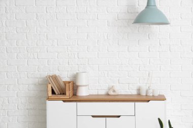 Odadaki beyaz tuğla duvarın yanında kitap ve vazoların olduğu çekmece.