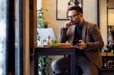 Handsome young businessman with glasses, using a laptop and a smart phone while working in the cafe