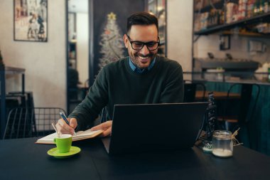 Smiling young man in eyeglasses writing in notebook while sitting in cafe