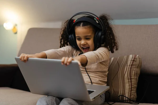 那个戴耳机的快乐的小女孩坐在沙发上 手里拿着笔记本电脑 — 图库照片