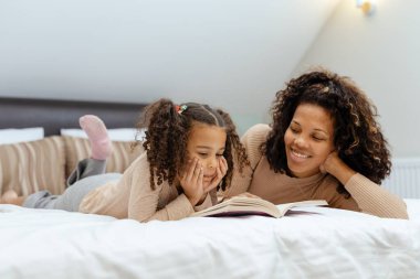 Evde yatakta uzanmış kitap okuyan anne ve kızın portresi.