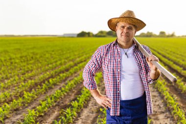 Yeşillik bir tarlada güvenle duran çiftçi omzunda çapa tutarak çok çalışmayı, tarımı ve kırsal yaşamı sembolize ediyor..