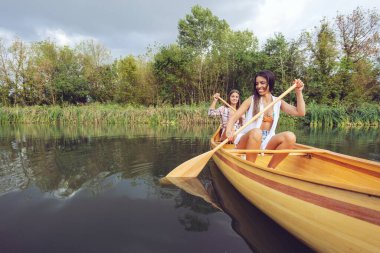 İki kadın, yemyeşil ve dingin doğayla çevrili sakin sularda, ahşap bir kanoda kürek çekerek huzurlu bir günün tadını çıkarıyorlar..
