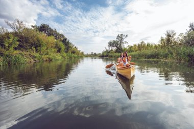 Bir insan, sudaki yansımaları ve bulutlu gökyüzü ile yemyeşil ve ağaçlarla çevrili sakin bir nehirde ahşap bir kanoda kürek çekiyor..