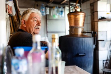 Yaşlı bir adam, çeşitli şişeler ve ahşap mobilyalarla çevrili klasik bir damıtma tesisi ile bir köy atölyesinde oturuyor..