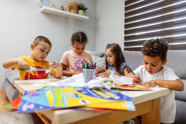 Dört çocuk bir masanın etrafında toplanıyor, kağıt ve renkli kalemlerle yaratıcı aktiviteler yapıyorlar, işbirliği ve hayal gücünü teşvik ediyorlar..