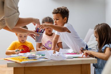 Çeşitli bir grup çocuk, parlak, rahat bir odada, bir yetişkinin rehberliği altında yaratıcılık ve takım çalışmasını ifade eden pastel boyalarla çizimlere odaklanıyor..