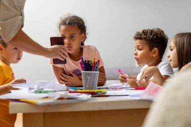 Çeşitli bir grup çocuk bir masanın etrafında oturur ve bir sanat etkinliğine katılırlar. Konferansta, bir yetişkinin rehberliğinde renkli malzemelerle el sanatları üzerinde duruluyor..