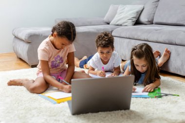 Üç çocuk rehberlik için dizüstü bilgisayar kullanarak görev yazmaya odaklanmış şekilde yerde yatıyor. Samimi bir oturma odasında öğrenme aktivitesi içindeler..