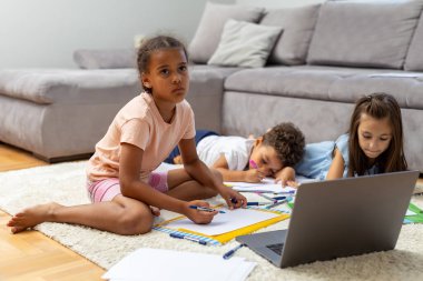Üç çocuk rahat bir ev ortamında yaratıcılık, öğrenme ve teknolojiyi gösteren rahat bir halının üzerinde otururken bir dizüstü bilgisayar çiziyor ve onunla meşgul oluyorlar..