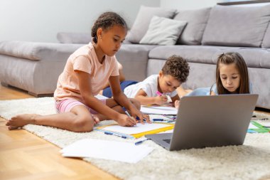 Üç çocuk rahat bir oturma odasında dizüstü bilgisayar çizmeye ve kullanmaya odaklanmış durumda. Rahat bir evde yaratıcılığı ve öğrenmeyi teşvik ediyorlar..