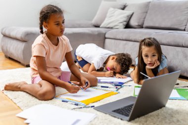 Üç çocuk bir çalışma oturumunda, modern bir oturma odasında yerde oturuyor, bir dizüstü bilgisayar kullanıyor ve uzaktan öğrenme aktiviteleri için materyaller çiziyor..