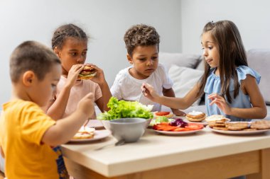 Dört çocuk bir masanın etrafında toplanıyor, taze sebzeli ve yayılmış sandviçleri birleştiriyor, takım çalışmasını ve sağlıklı yeme alışkanlıklarını sıcacık bir ortamda ön plana çıkarıyor..