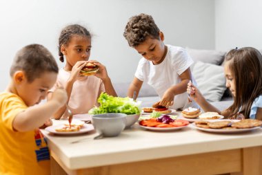 Dört çocuk bir masada oturup hamburger, salata ve diğer yemekleri yiyor, birlikte yemek yiyor, sohbet ediyor ve zamanlarının tadını çıkarıyor..