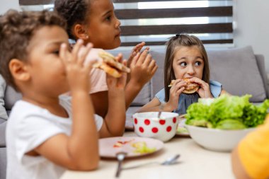 Üç çocuk bir masada hevesle hamburger yiyorlar. Masa taze marulla döşenmiş, hayat dolu, rahat ev düzenine biraz yeşillik katılmış..