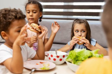 Üç çocuk bir masada oturmuş ortada salata kasesiyle hamburgerlerin tadını çıkarıyorlar. Sahne, birlikte geçirdiğimiz gündelik ve mutlu yemek zamanlarını tasvir ediyor..