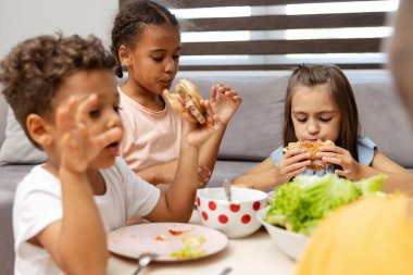 Üç çocuk parlak bir masada toplanmış, mutlu bir şekilde hamburger ve diğer yiyecekleri yiyorlar, modern bir evde neşeli ve sıradan bir yemek sergiliyorlar..