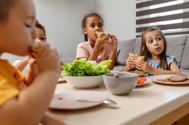 Bir grup çocuk bir masanın etrafında oturmuş, sıradan bir yemeği paylaşıyorlar. Sahne, rahat bir evde yemek yiyen ve etkileşime geçen çocuklarla dolu..