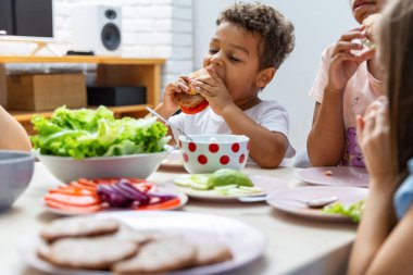 Genç bir çocuk renkli taze sebze tabaklarıyla çevrili ev yapımı burgerden büyük bir ısırık alıyor. Bu da evde eğlenceli ve sağlıklı bir yemek zamanı olduğunu gösteriyor..