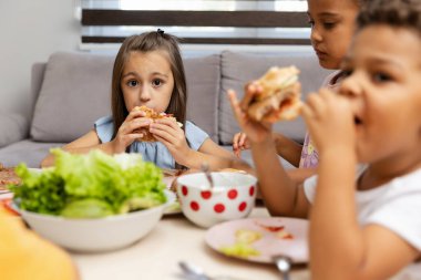 Üç çocuk bir masada oturmuş taze sebzelerle çevrili sandviçlerini yiyor ve rahat evlerinde rahat bir yemek yiyorlar..