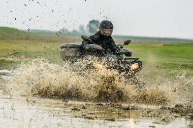 ATV 'deki bir sürücü açık alandaki çamurlu su birikintilerinde güçlü bir şekilde şarj oluyor, çamur ve su sıçrıyor, macera sergiliyor ve heyecan içinde hareket ediyor..