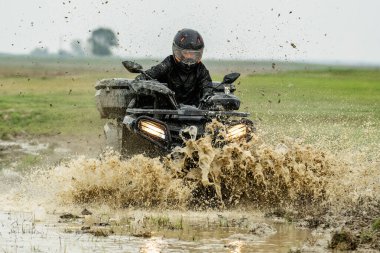 Koruyucu giysili bir insan çamurlu bir su sıçramasında ATV kullanır, hareketli ve hareketli bir sahnede off-road yolculuğunun heyecanını ve macerasını yakalar..