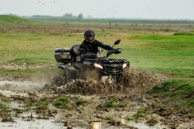 ATV 'deki bir kişi çamurlu bir arazide geziniyor, dinamik bir çamur ve su sıçramasına neden oluyor, açık ve otlak bir arazide heyecan ve macera sergiliyor..