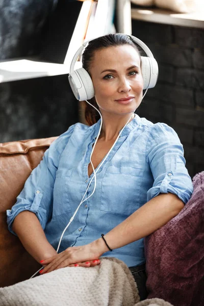 Leisure time concept. Happy beautiful woman listening to the music using headphones sitting on a couch indoors. Female spending her free day and relaxing at home alone