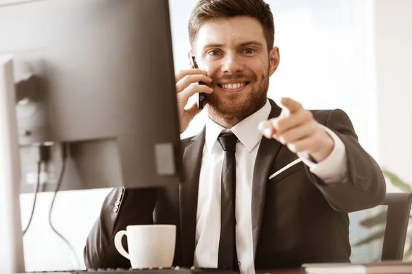 stock image Business concept. Happy smiling young businessman sitting in office talking on a cell phone getting good news about his work and pointing with finger. Man in suit indoors on glass window background.