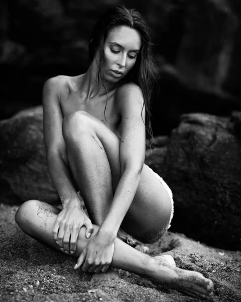 夏季娱乐的概念 美丽的性感女子 身材匀称 训练有素 身材苗条 穿着蓝色短裤 坐在石头上的海滩上 时尚女模特在海边摆姿势 黑白单色 — 图库照片
