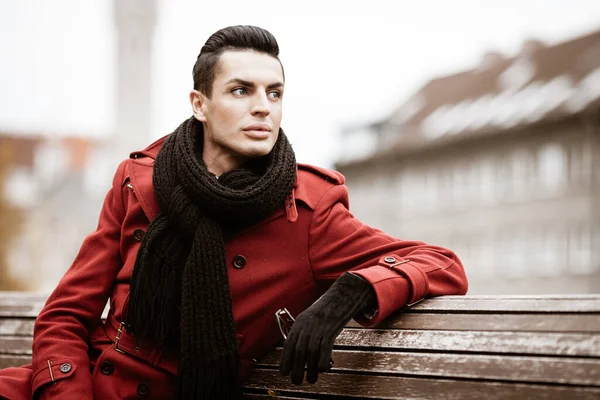 LGBTQ community lifestyle concept. Young homosexual man sits on bench in city park. Handsome fashionable gay male model poses in cityscape outdoors. Wears red coat, gloves, and black scarf.