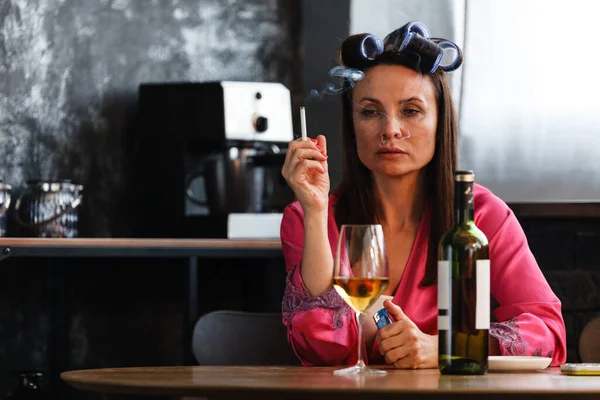 Alcoholism problem concept. Sad middle aged woman in stress sits in the kitchen, drinks white wine from a glass and smokes a cigarette. A depressed female is addicted to alcohol at home