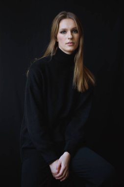 Siyah kıyafetli, esmer arka planda oturan genç bir kadının zamansız portresi. Yumuşak ışıklandırma onun yüz hatlarını ve zarif duruşunu vurgular, klasik ve sofistike bir görünüm yaratır..