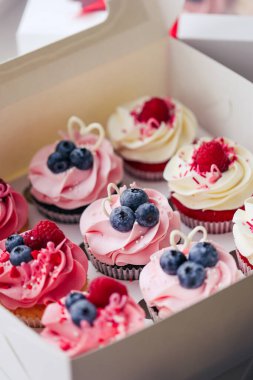 Fresh and delicious cupcakes with cheese cream and fresh berries. Top view of arranged tasty cupcakes in cardboard box isolated on white. Cakes with cream, chocolate bars, blueberries and raspberries
