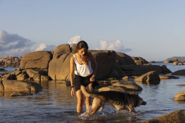 Plajda gün batımında köpekle oynayan genç Latin kadın.