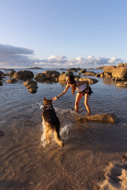 Plajda gün batımında köpekle oynayan genç Latin kadın.