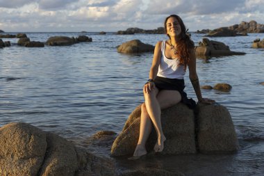 Deniz kenarındaki genç Galiçyalı kadın gün batımında okyanusun yanındaki bir kayanın üzerinde oturuyor.