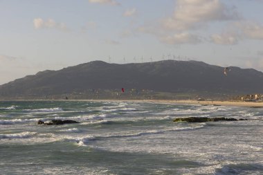 Parlak güneşli bir günde, Tarifa sahilinde bulutlarla sörf yapan birçok insan uçurtma sörfü yapar..