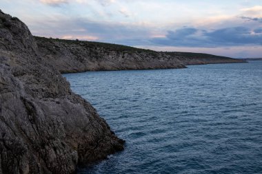 Alacakaranlıkta kayalık bir sahil şeridinden nefes kesici manzara, dingin sulardaki ışık ve gölgenin etkileşimini ve engebeli manzarayı yakalıyor..