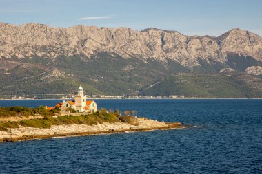 Bu büyüleyici görüntü, arka planda deniz güzelliği ve maceranın cazibesini somutlaştıran resimli dağlarla parıldayan mavi suların yanında bir deniz feneri barındırıyor..
