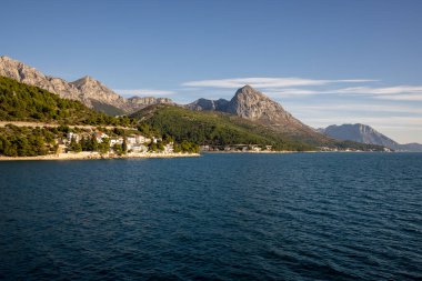 Nefes kesici bir manzara, sakin mavi suların yanında görkemli dağlar sergiliyor, doğanın büyüleyici güzelliğini ve bu kıyı manzarasının sükunetini vurguluyor..