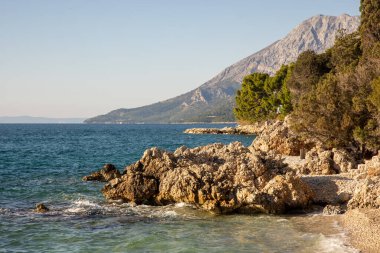 Kayalık sahilleri, berrak turkuaz suları ve arka planda uzanan görkemli bir dağı gösteren sakin bir sahil sahnesi rahatlama ve keşifleri davet ediyor..